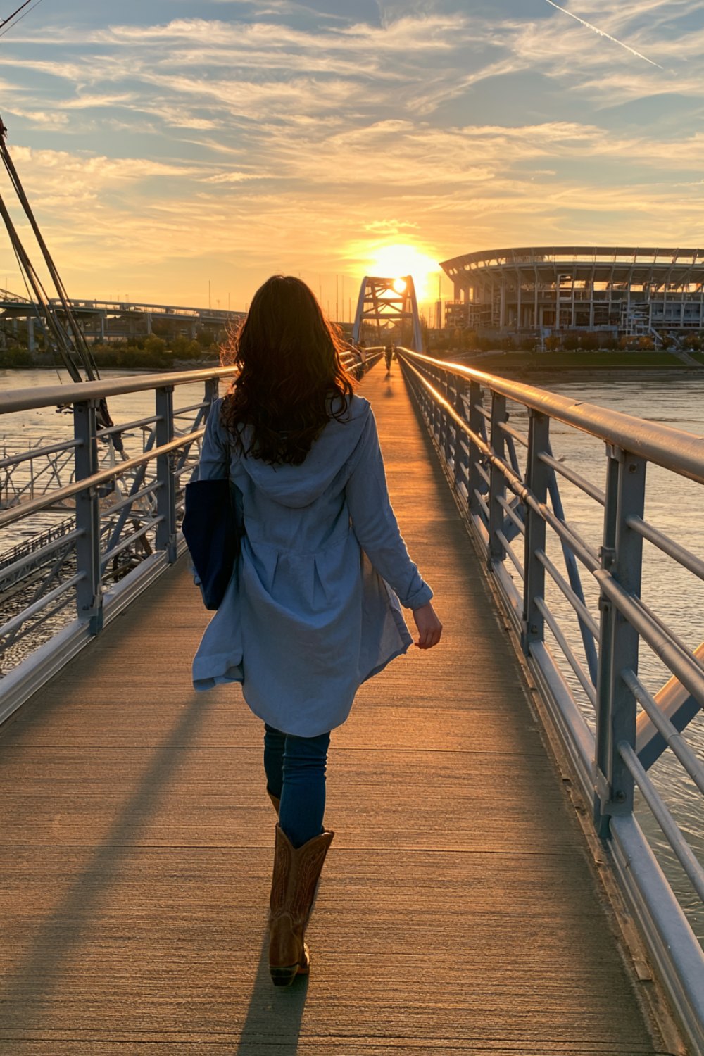 Woman from behind in light blue and navy walking a wooden bridge toward a stadium at sunset