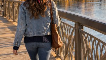 Woman seen from behind on a riverside pedestrian bridge in a light blue denim jacket, red bandana and brown cowboy boots, with a stadium ahead