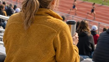 Mom seen from behind on bleachers at an outdoor track meet in a mustard fleece and cap holding a travel mug