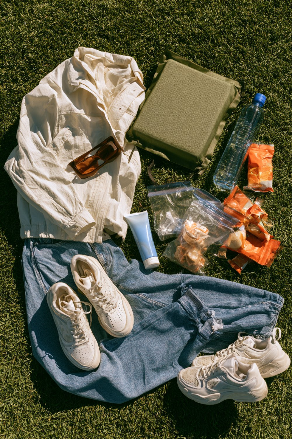 Track meet day kit on grass: jacket, jeans, sneakers, sunscreen, stadium cushion and snacks