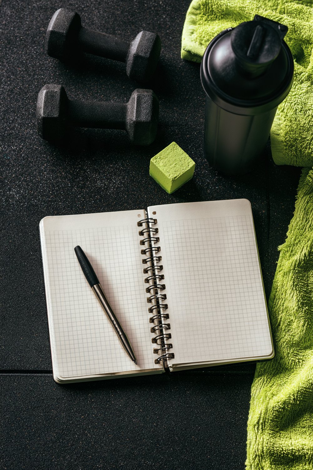 Open training notebook with a pen next to dumbbells, a shaker bottle and a towel on a rubber gym floor