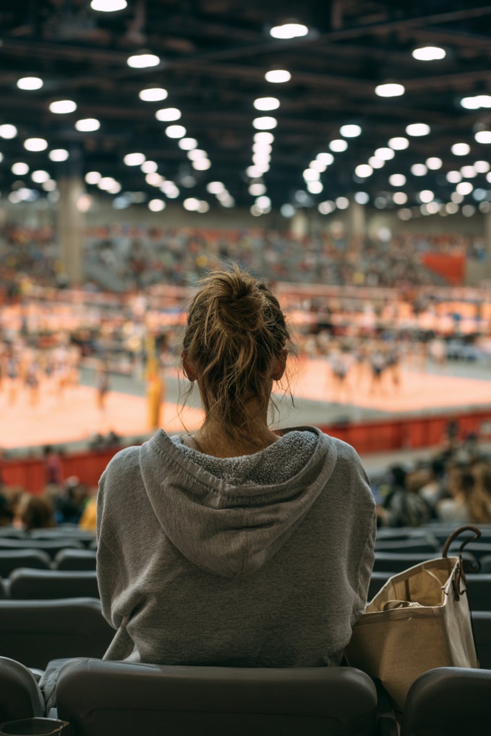 Woman in a grey hoodie seen from behind in the stands of a large indoor volleyball tournament, another angle
