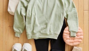 Flat lay on a maple gym floor of a sage crewneck, black flared leggings, white sneakers, tote bag and pink tumbler