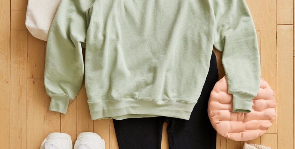 Flat lay on a maple gym floor of a sage crewneck, black flared leggings, white sneakers, tote bag and pink tumbler