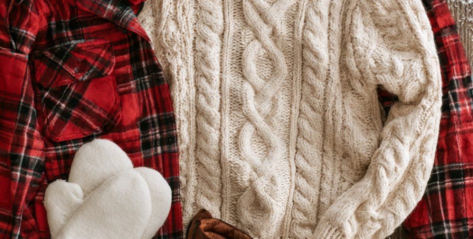 Flat lay on a snowy table of a red plaid flannel, cream cable knit sweater, red pom beanie, mittens and a thermos