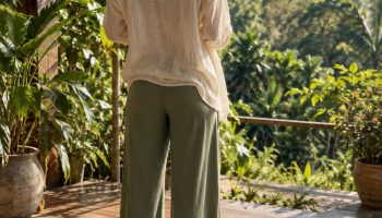 Woman seen from behind on a yoga mat on a sunlit deck in a loose cream linen top and sage wide leg yoga pants